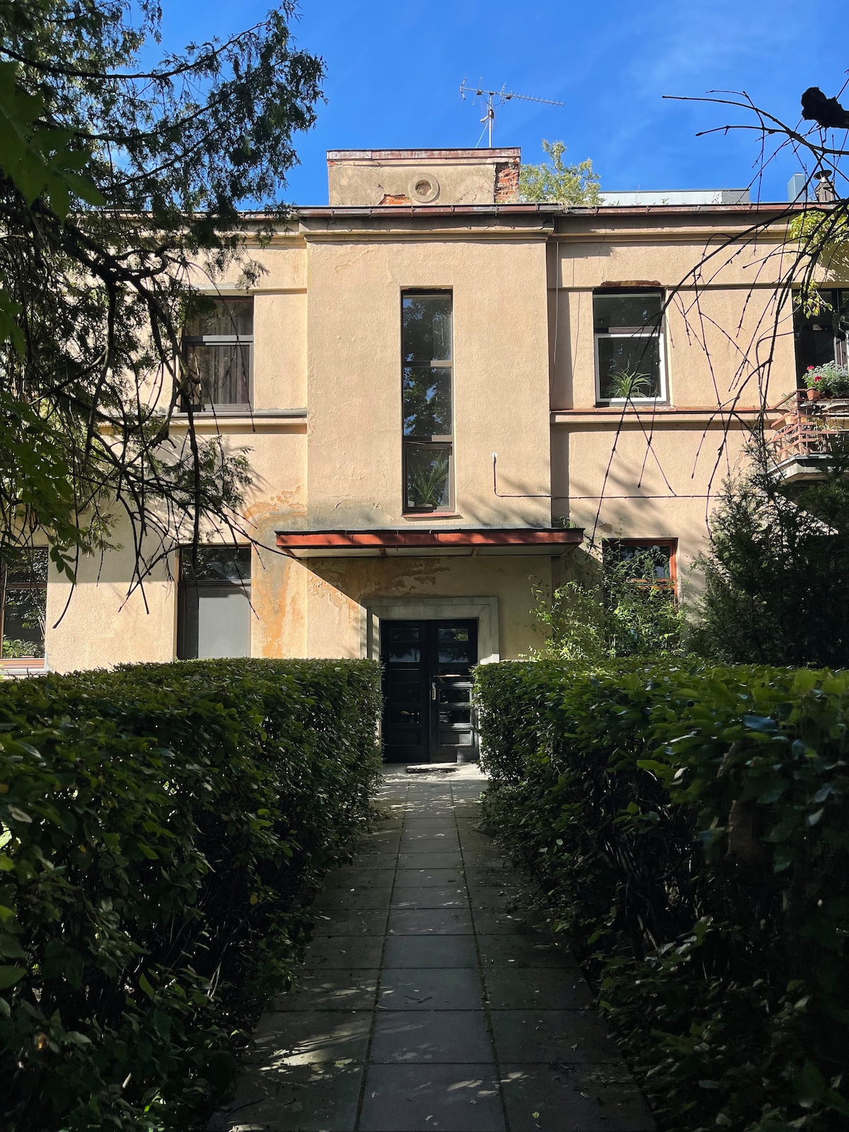 The entrance of the apartment building is framed by well-maintained hedges and a stone pathway leading to the door. Sunlight filters through the trees, illuminating the building's vintage façade and highlighting large windows.