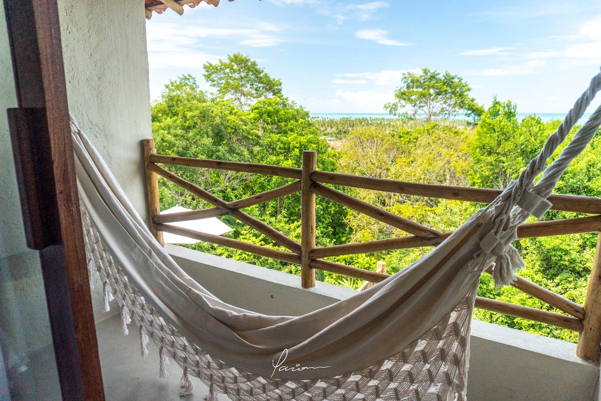 A spacious balcony is shown, featuring a comfortable hammock suspended between wooden posts. Lush greenery is visible beyond the railing, with a glimpse of blue water in the distance. The clear sky adds to the serene atmosphere, inviting relaxation.