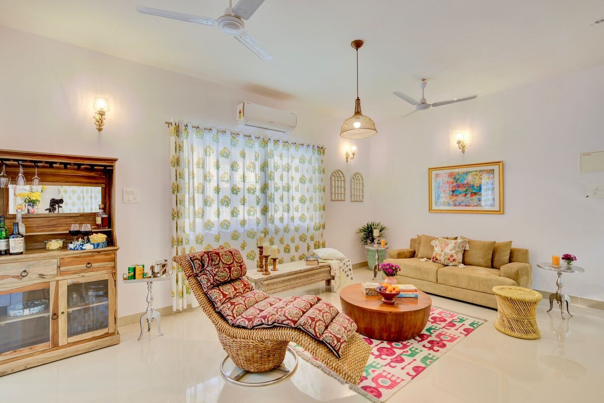 The living area is furnished with a comfortable sofa, a woven chair, and a round coffee table. Decorative elements include a vibrant area rug and colorful artwork on the wall. Natural light filters through sheer curtains adorned with floral patterns.