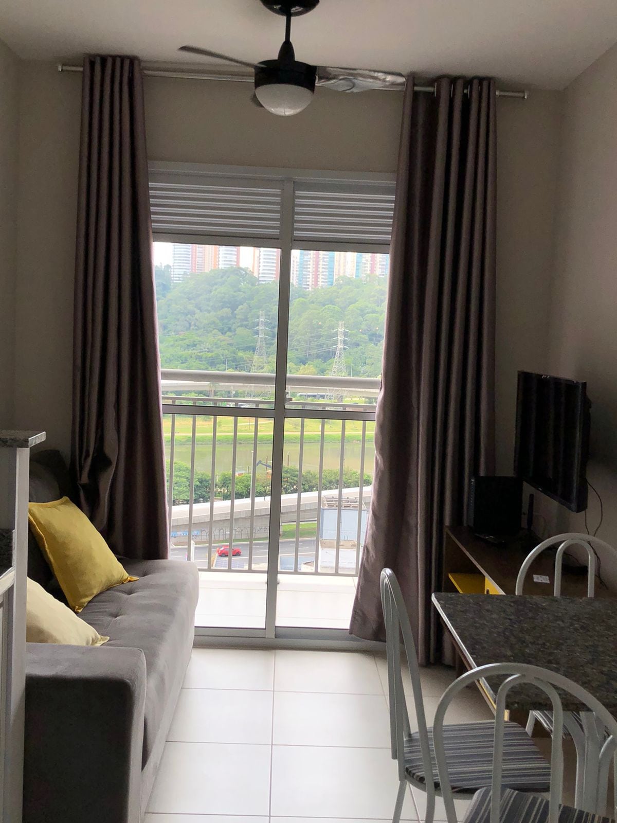 A cozy sitting area features a gray sofa with yellow pillows and a flat-screen TV. Large glass doors provide access to a balcony, offering views of greenery and nearby buildings. Natural light brightens the room, complemented by neutral-toned curtains.