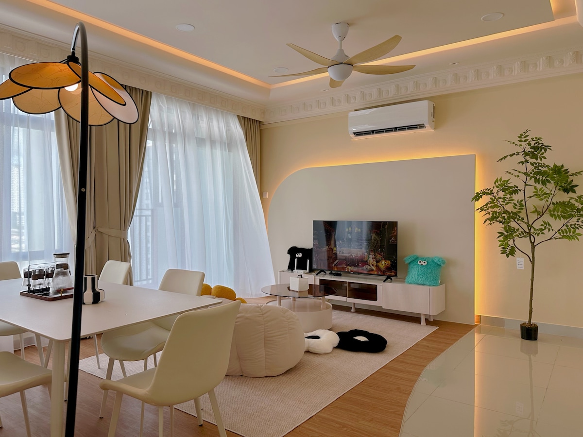 The living area features large windows dressed in sheer curtains, allowing natural light to fill the space. A comfortable seating arrangement is present, including a modern sofa and floor cushions. A dining table for six is positioned nearby, and a wall-mounted television is visibly placed on a console.