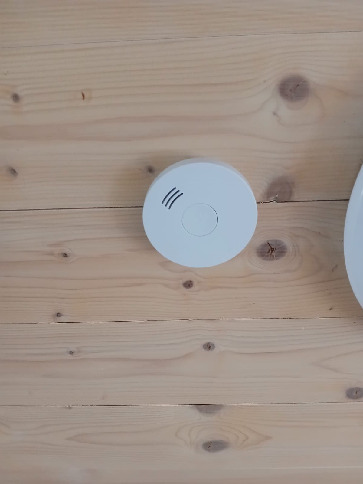 A smoke detector is mounted on a light wooden ceiling. The detector is round and white, featuring a small button in the center and three horizontal lines above it. The natural wood grain is visible around the detector, adding a warm touch to the setting.