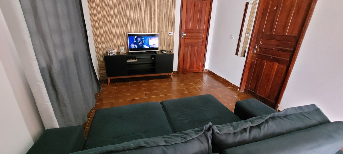 A living area is presented with a dark sofa facing a television set. A wooden cabinet holds the TV, and natural light enters through a nearby window with grey curtains. The back wall is textured, adding a subtle detail.