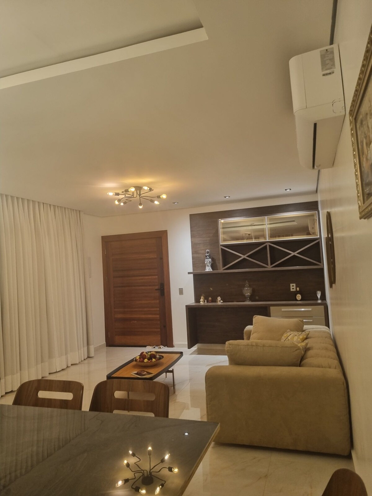 A modern living area features a comfortable beige sofa and a wooden coffee table. Light-colored curtains frame the window, allowing natural light to enter. A wooden cabinet with glass shelving is positioned against the wall, enhancing the space's functionality.