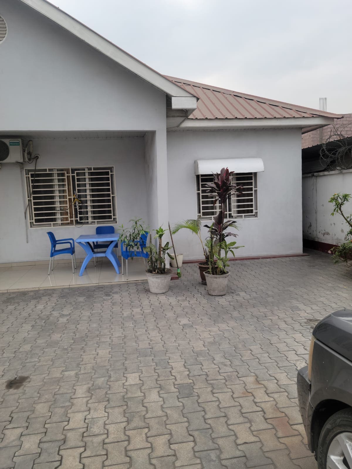A light grey exterior of a house is presented, featuring a small seating area with blue chairs on a tiled patio. Two potted plants are positioned beside the entrance, with a window adorned by a white awning. The pathway is paved, enhancing the property's inviting ambiance.