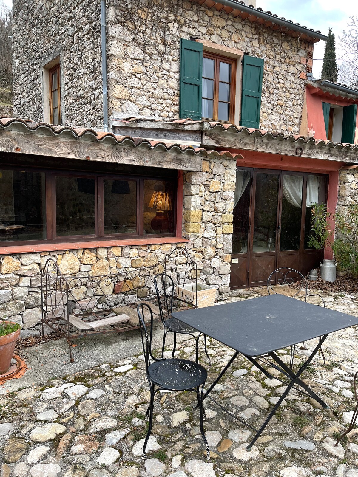 An outdoor seating area is set on a stone patio, featuring a metal table surrounded by four chairs. Stone walls provide texture to the building, while green shutters frame large windows that invite natural light into the space.
