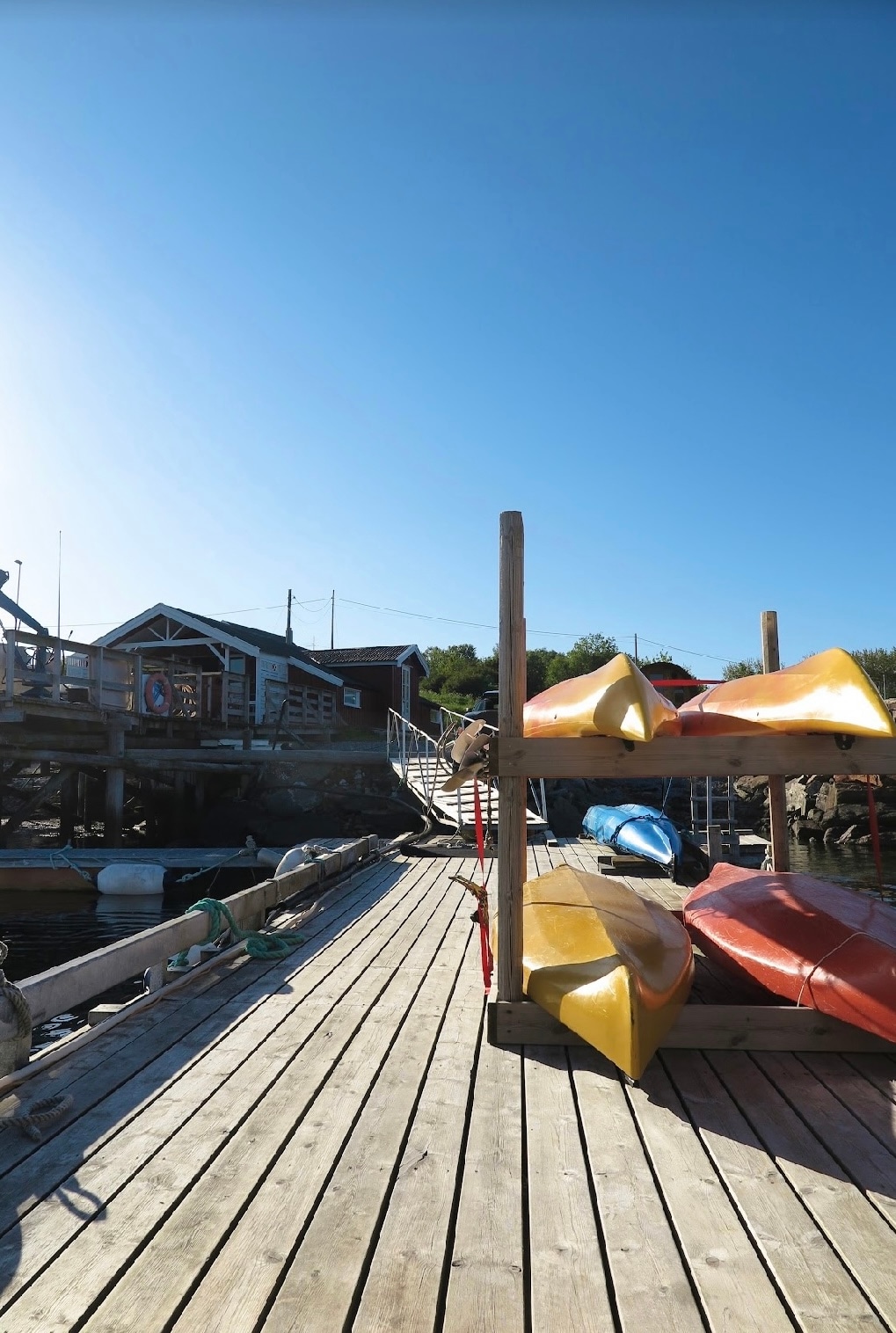 A wooden dock is visible, lined with brightly colored kayaks in shades of yellow, orange, and blue. A building can be seen in the background, with steps leading up from the dock. The scene is set against a clear blue sky, suggesting a serene outdoor environment.