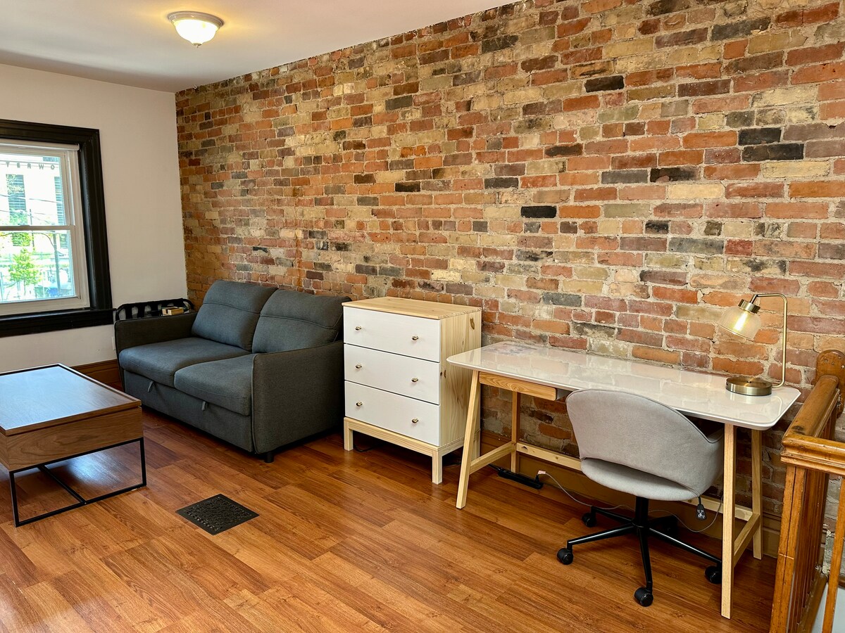 A cozy sitting room features a grey sofa and a modern wooden coffee table, complemented by a white dresser and a sleek desk with a chair. Exposed brick walls add warmth, while natural light filters in through the nearby window.