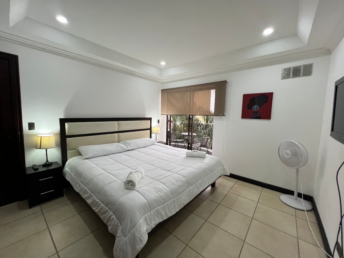 A spacious bedroom is furnished with a large bed, complemented by a bedside lamp and nightstand. Natural light flows in from large windows covered with shades. A decorative wall piece adds a touch of character, and a fan provides additional comfort.