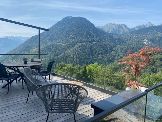 A spacious terrace features a wooden floor and sleek glass railing, offering panoramic views of the surrounding mountains. A round dining table is accompanied by four chairs, while two stylish armchairs provide additional seating, creating an inviting outdoor area for relaxation.