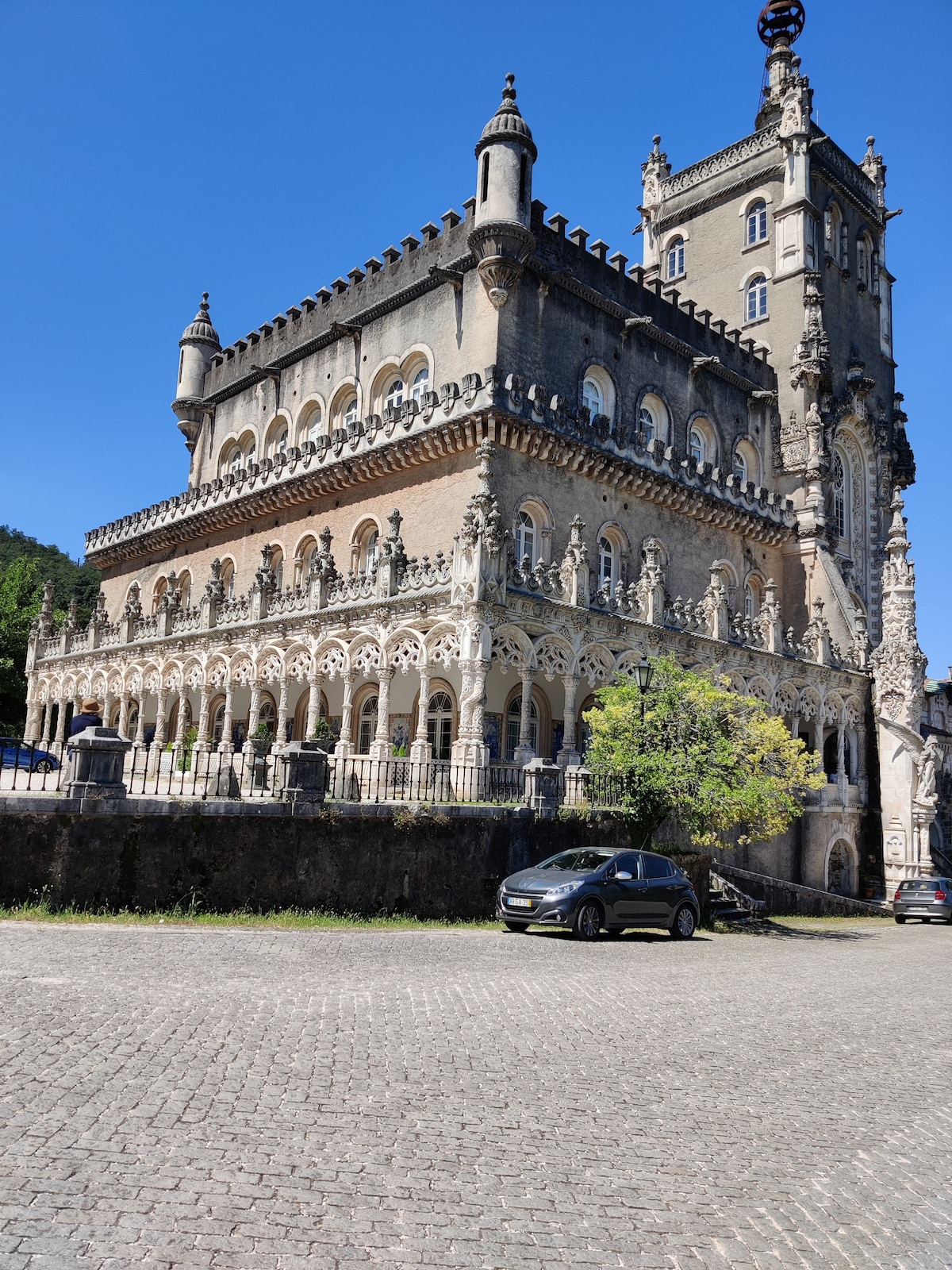 A grand historical building features intricate architectural detailing with tall towers and ornate balconies. The structure is set against a clear blue sky, surrounded by well-maintained grounds. A parked car can be seen in the foreground, providing a sense of scale to the impressive facade.