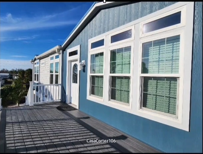 The exterior of the stilt home features a blue facade with multiple large windows, allowing for natural light. A white railing lines the small porch, which leads to the entrance. The surrounding area includes greenery, enhancing the inviting appearance.