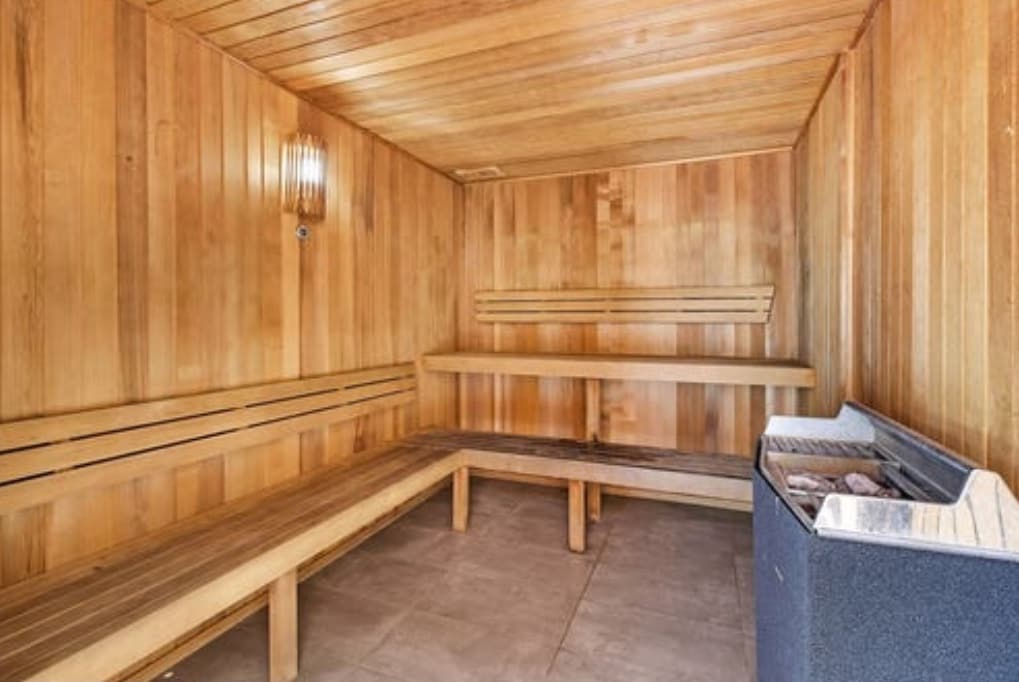 A wooden sauna features lined benches along the walls, providing seating for multiple guests. Soft lighting illuminates the warm wooden interiors, creating a serene atmosphere. A stone heater is positioned against one wall, designed for a calming sauna experience.