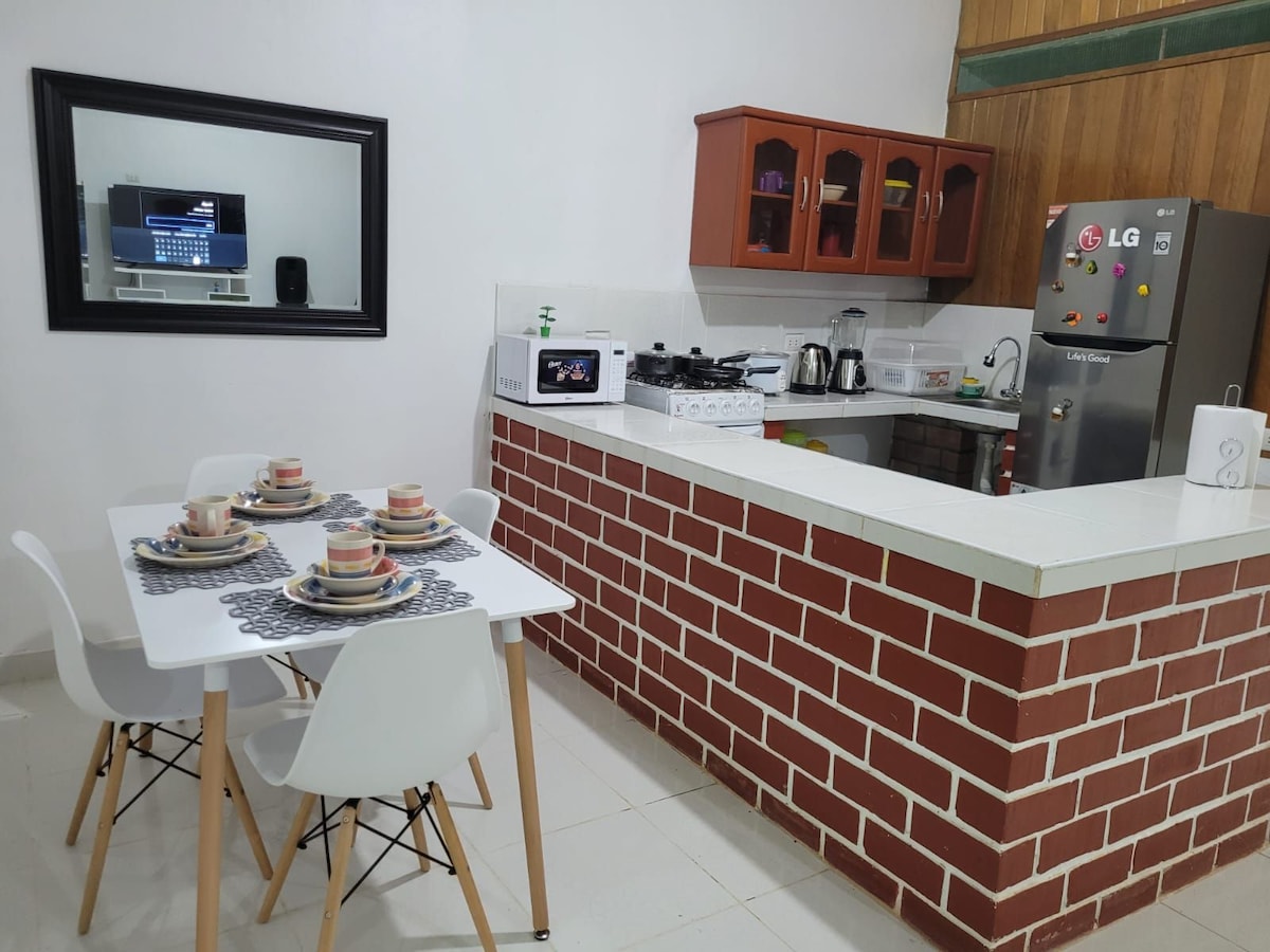 A bright kitchen area features a modern stove and refrigerator against a backdrop of wooden accents. A dining table is set for four, with patterned plates and cups arranged on woven placemats. A large mirror reflects the space, enhancing the feeling of openness.