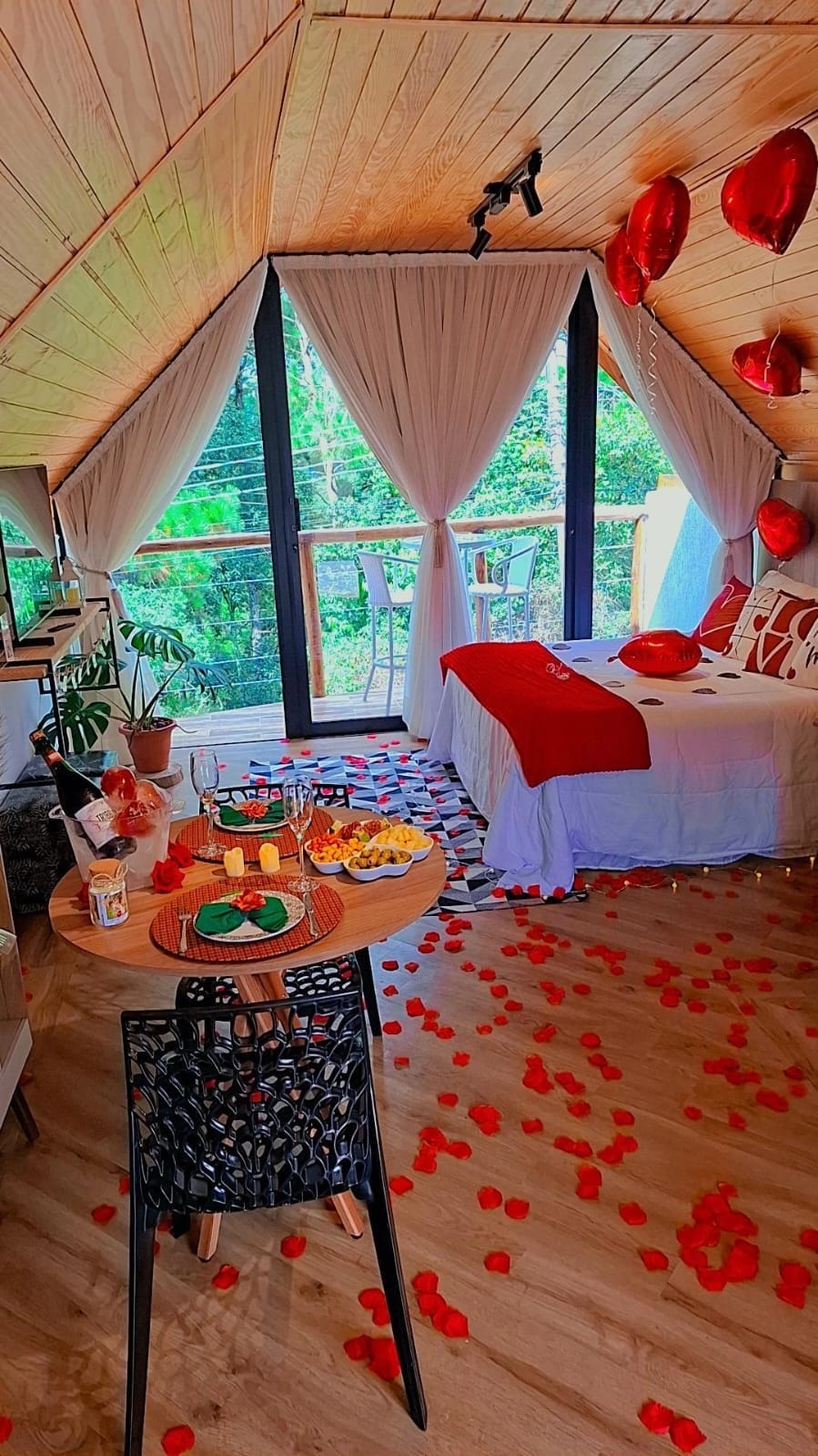 An inviting interior features a spacious sleeping area draped in red accents and decorative pillows. A small dining table is set for two, surrounded by scattered rose petals. Soft lighting enhances the atmosphere while large windows provide views of the lush greenery outside.