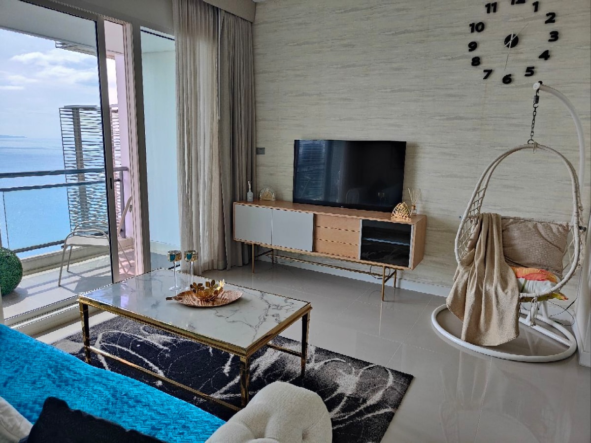 A modern living area is depicted, featuring a wall-mounted television above a stylish media console. A round swing chair offers a cozy seating option, complementing the marble coffee table and plush area rug. Natural light fills the space through large glass doors leading to the balcony.