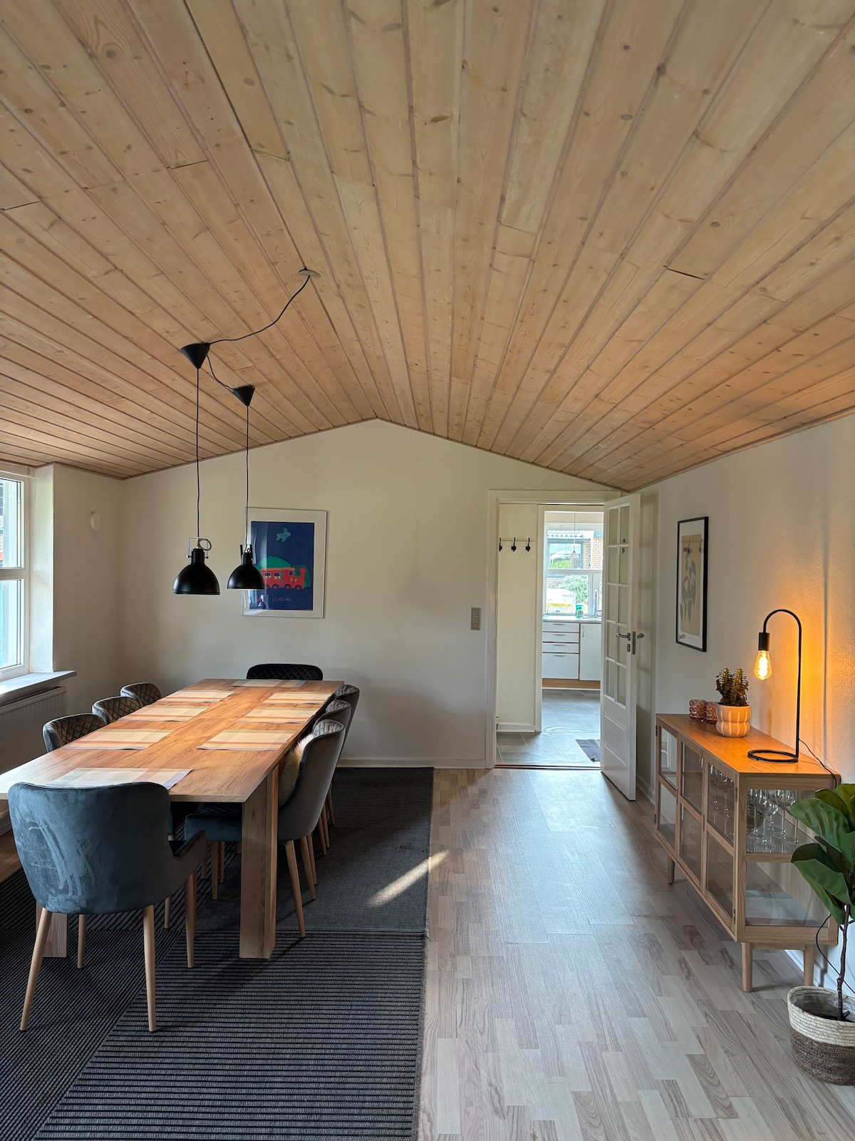 A bright dining area is featured with a long wooden table surrounded by upholstered chairs. Two black pendant lights hang above the table, and a warm floor lamp is positioned beside a minimalist side table. A door leads to another room, enhancing the open feel.