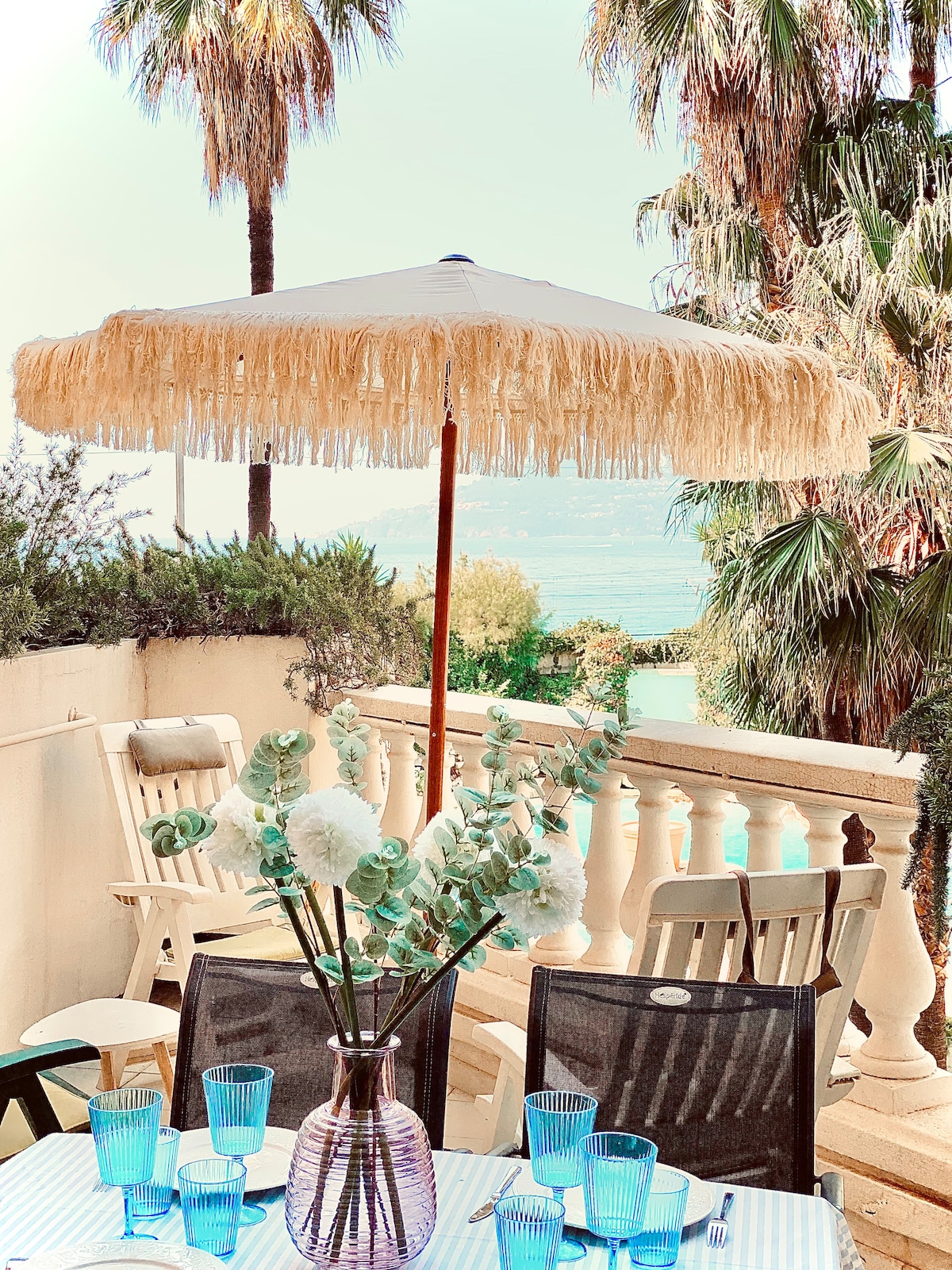 A terrace setting is presented with a dining table topped with glasses in soft blue tones. A vase filled with greenery and flowers adds elegance, while a fringed umbrella provides shade. In the background, palm trees and a glimpse of the water can be seen.