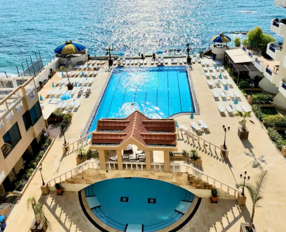 A large outdoor swimming pool is framed by sun loungers and umbrellas, with a waterfront view. The area is neatly arranged, featuring palm trees and landscaped greenery. Sunlight reflects off the water, creating a shimmering effect, while spacious seating areas are strategically placed around the pool.