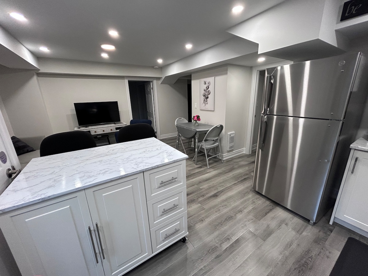 A modern kitchen area with a marble-topped island and white cabinetry is highlighted. Stainless steel appliances, including a refrigerator, complement the space. A small dining table with folding chairs can be seen, while soft lighting enhances the inviting atmosphere.