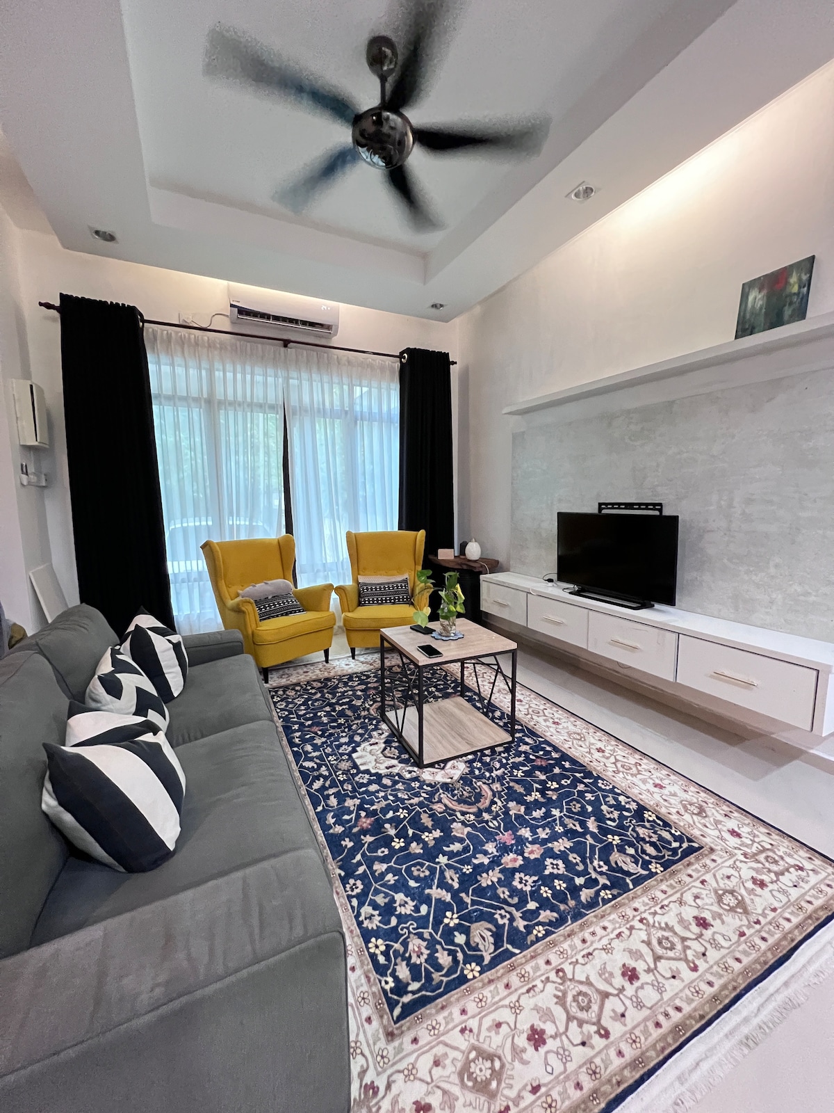 A modern living area features gray sofas with striped cushions and two yellow armchairs. A coffee table sits centrally on a patterned rug. A television stands on a white console against a textured wall, with large windows dressed in dark curtains allowing natural light to filter in.