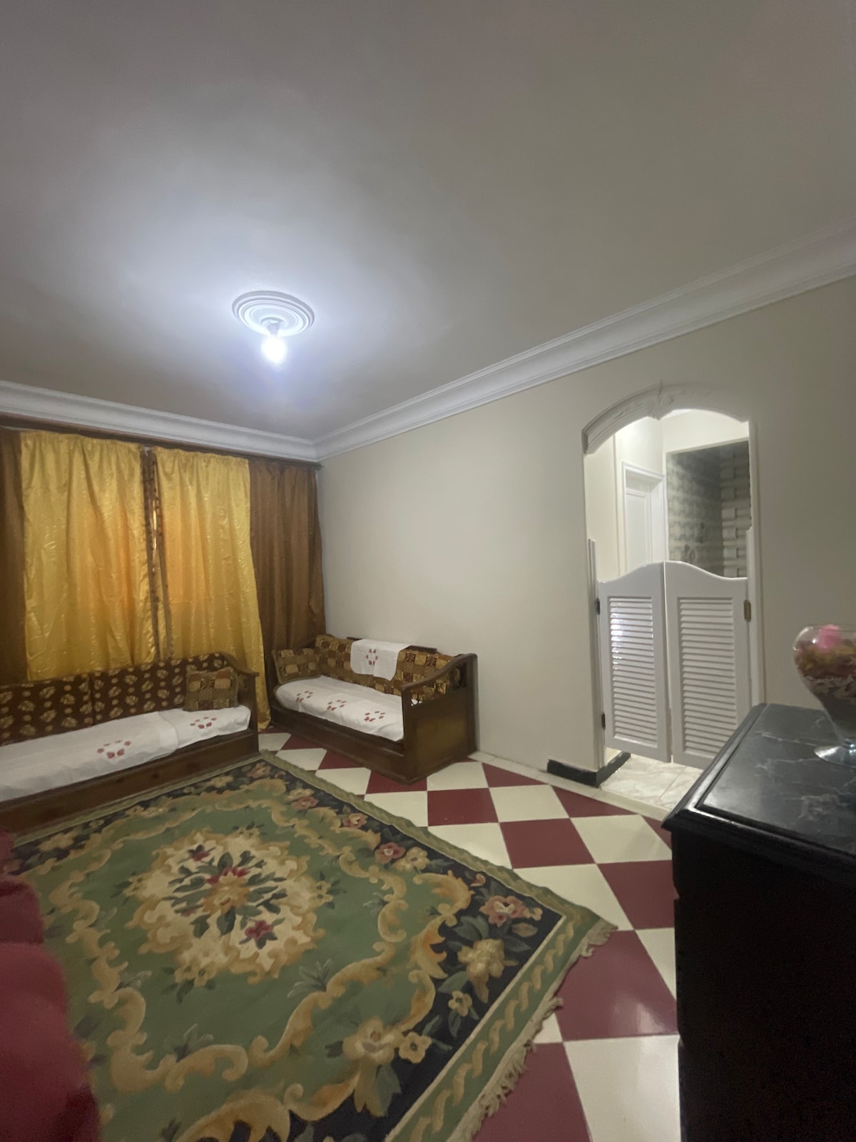A well-appointed living area features patterned tiles and a large area rug. Two wooden sofas are positioned against a wall, accented by decorative cushions. Natural light filters through sheer curtains, illuminating the space. An archway leads to an adjoining room, enhancing the home's open feel.