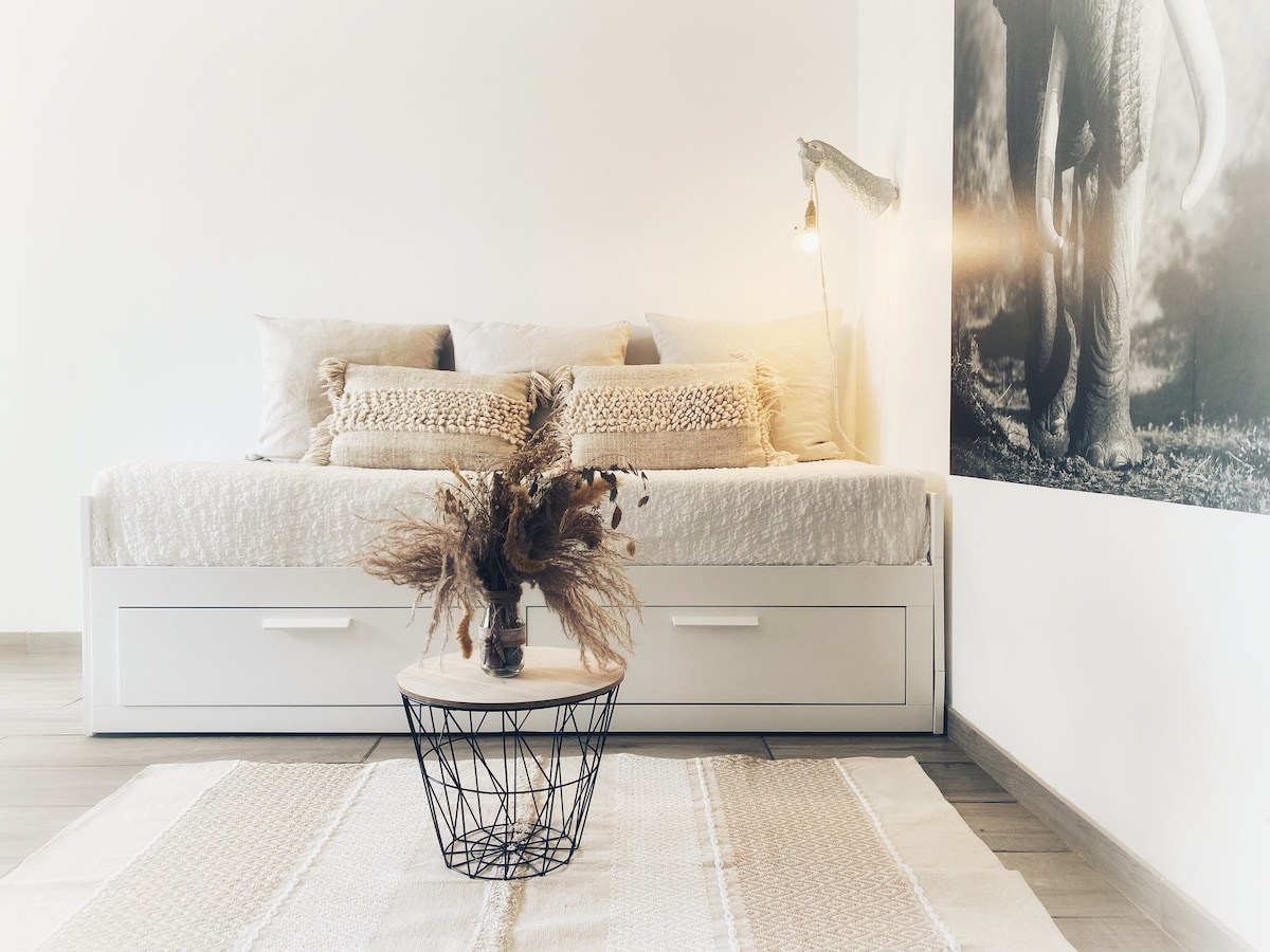 A cozy sleeping area features a spacious bed with decorative cushions. A small table with a wire design is placed at the center, adorned with a dried floral arrangement. A calming image of an elephant is displayed on the wall, complementing the serene atmosphere.