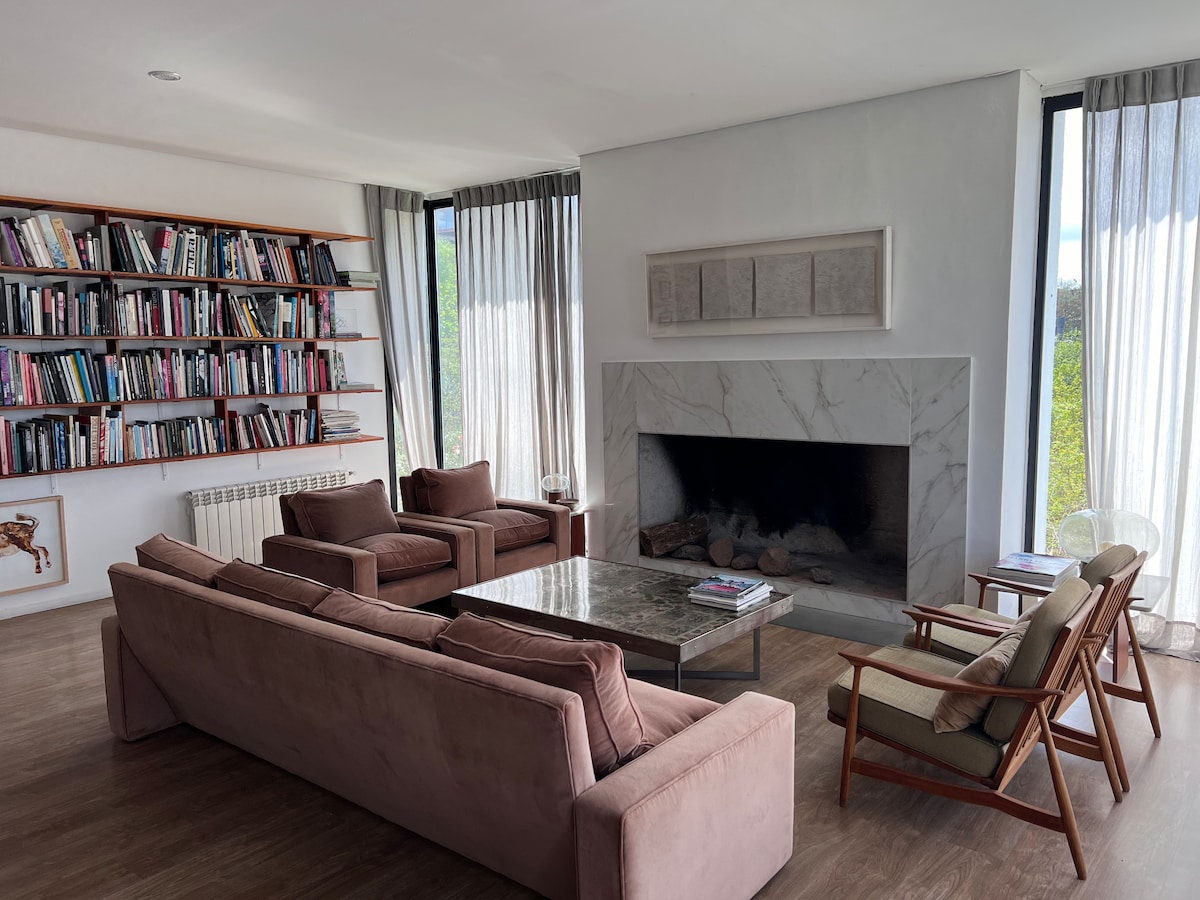 A spacious living area features a modern design with large windows allowing natural light. A stone fireplace is central to the space, with soft brown seating arranged around a marble coffee table. Shelves filled with books line one wall, enhancing the serene ambiance.