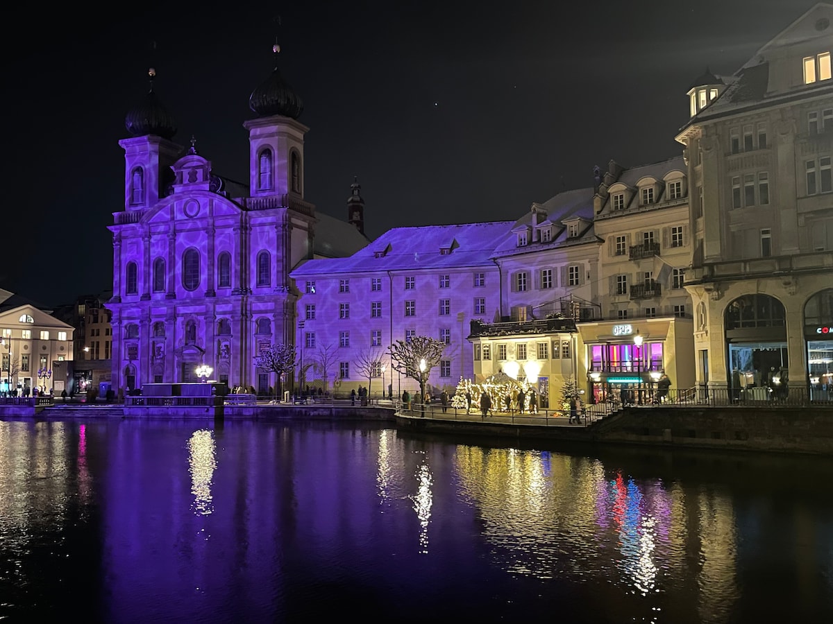 A scenic nighttime view of a historic building alongside a calm waterfront. Soft purple lighting highlights the architecture, while reflections shimmer on the water's surface. Various people are seen gathered outside a nearby establishment, contributing to the lively atmosphere of the area.