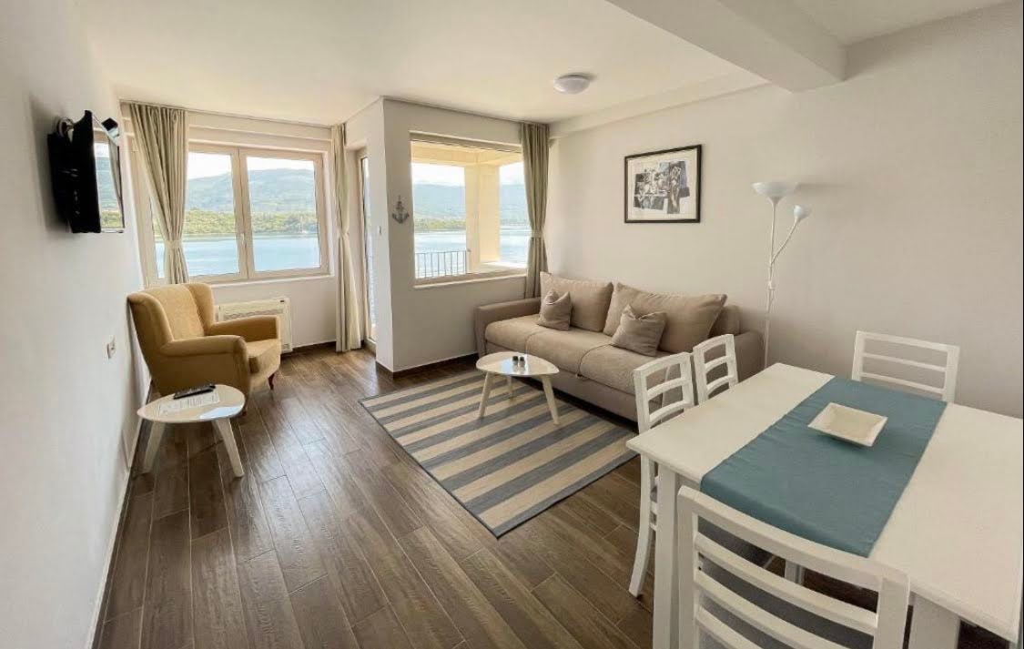 A bright living area features a comfortable sofa and a cheerful yellow armchair, complemented by a modern coffee table. Large windows provide natural light and scenic views of the nearby water. A dining table with white chairs is positioned beside a rug with striped patterns.