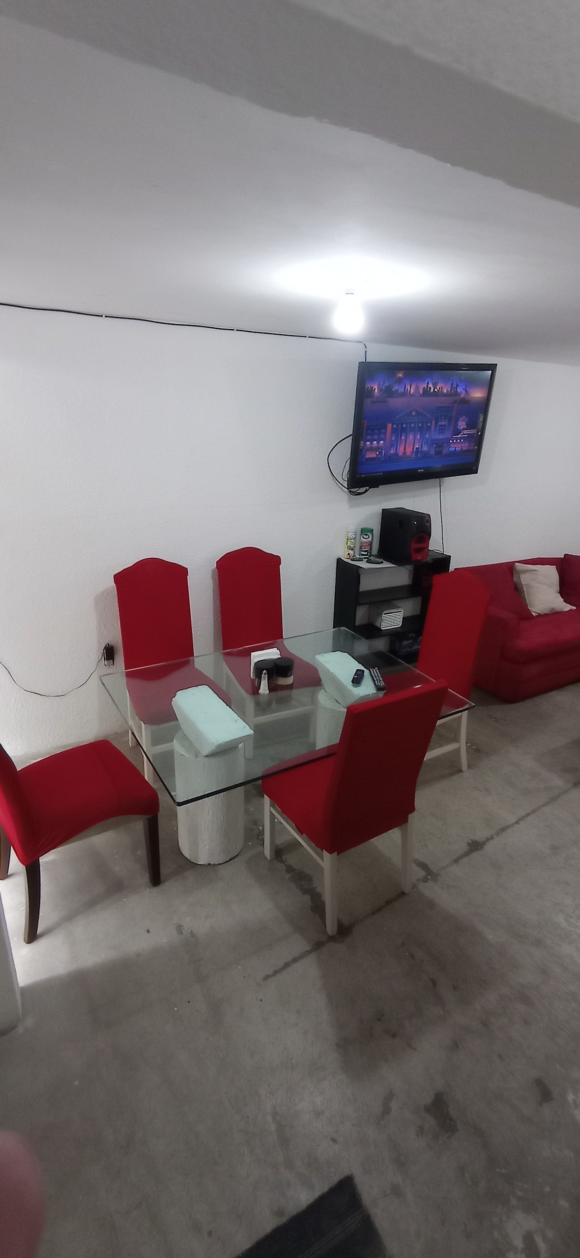 A dining area is presented with a glass table surrounded by six red chairs. A red sofa is positioned nearby, and a television mounted on the wall is clearly visible. Soft lighting is provided by a ceiling fixture, creating a functional and comfortable space.