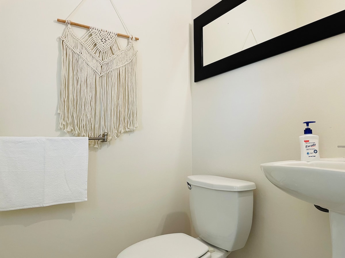 A neutral-toned bathroom features a white toilet and a small sink. Above the sink, a rectangular mirror reflects the simple design. A decorative macrame wall hanging provides a textural element, while a white hand towel is neatly displayed on a towel bar.
