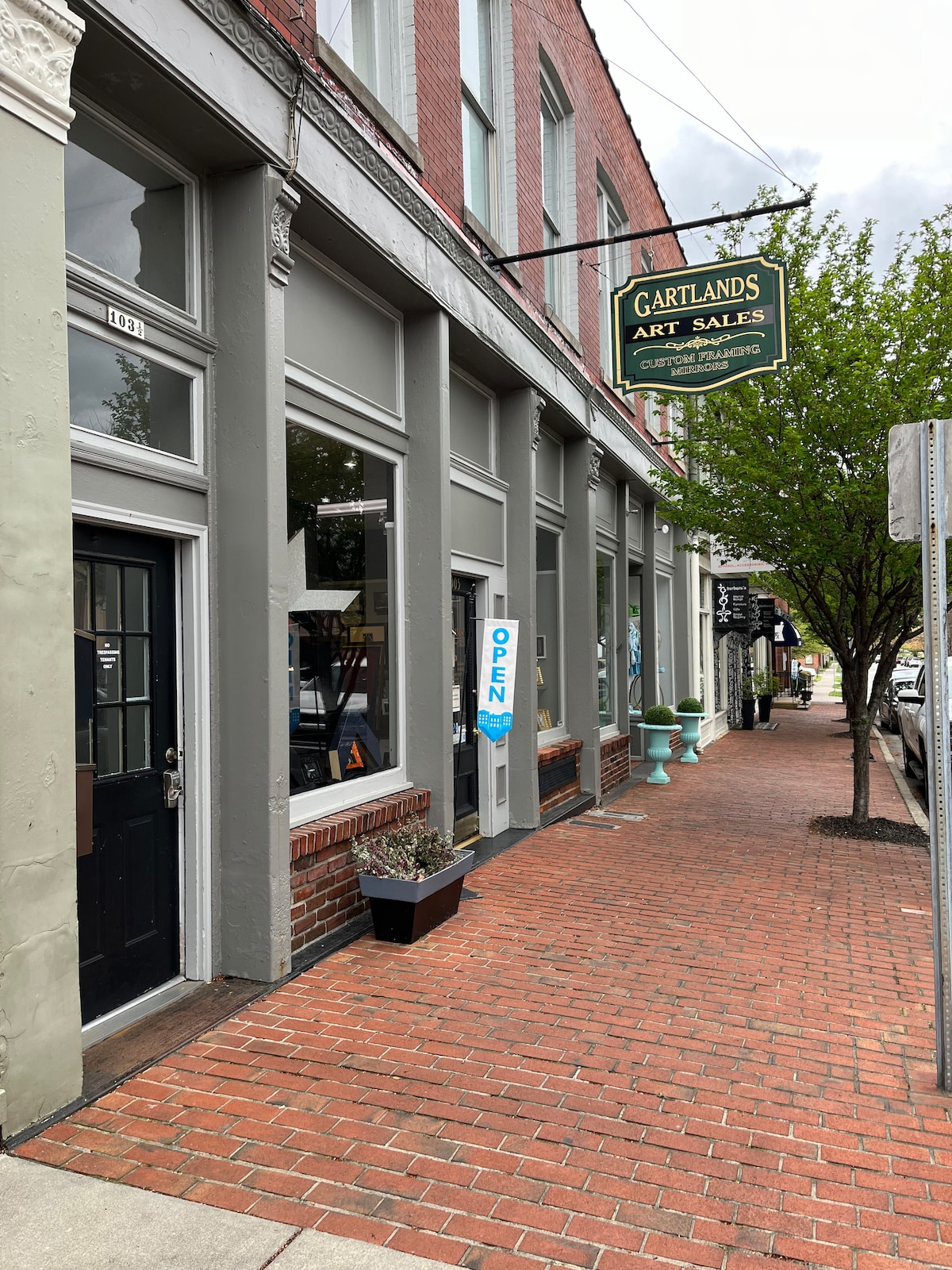 The image showcases the exterior of Gartlands Art Sales. A brick sidewalk leads to a storefront featuring large windows displaying art pieces. An 'OPEN' sign is visible in the door, and potted plants decorate the entrance, enhancing the welcoming atmosphere of the downtown area.