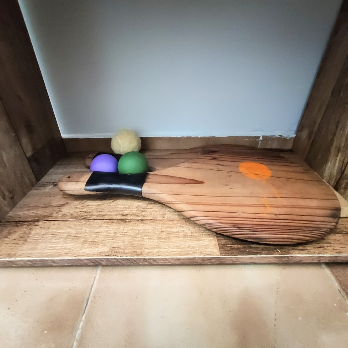 A wooden paddle is positioned upright on a wooden shelf. Beside it, three decorative balls in purple, green, and beige are arranged, adding a touch of color to the natural wood tones.
