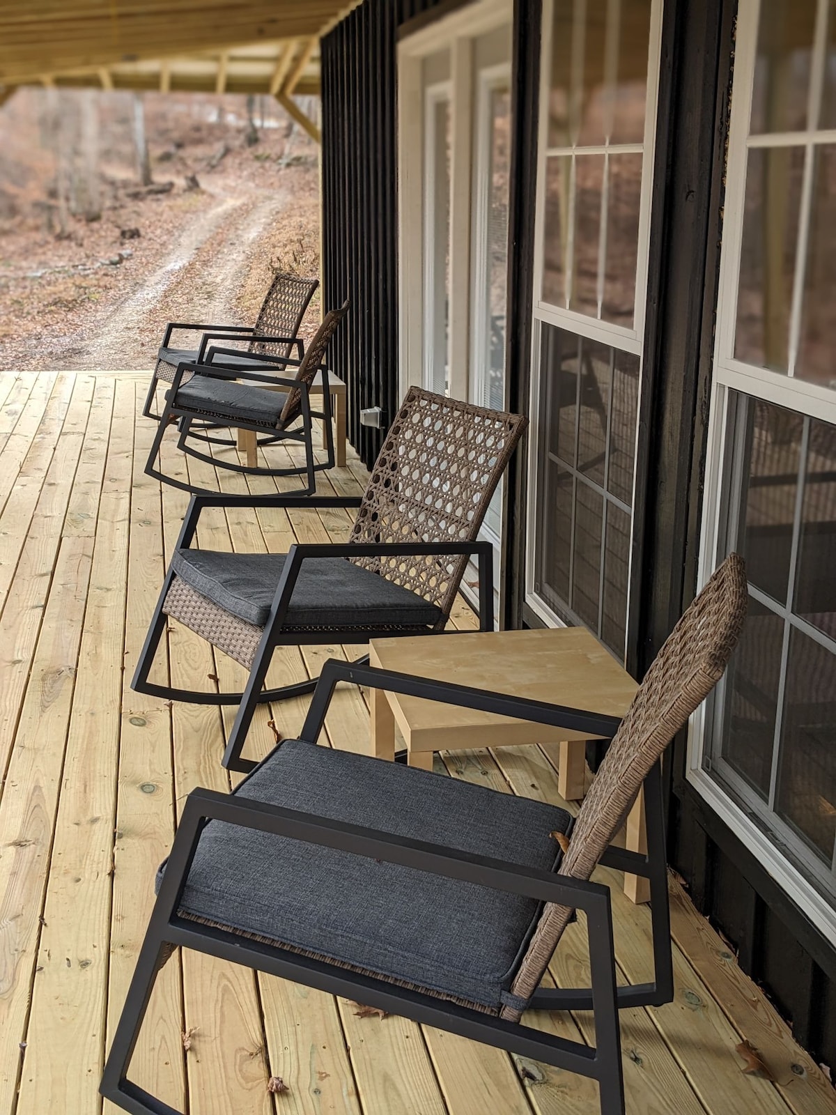A covered porch is furnished with several rocking chairs and a small side table. The chairs are designed for comfort and relaxation, placed on a wooden deck that leads to a natural setting, offering a view of the surrounding landscape.