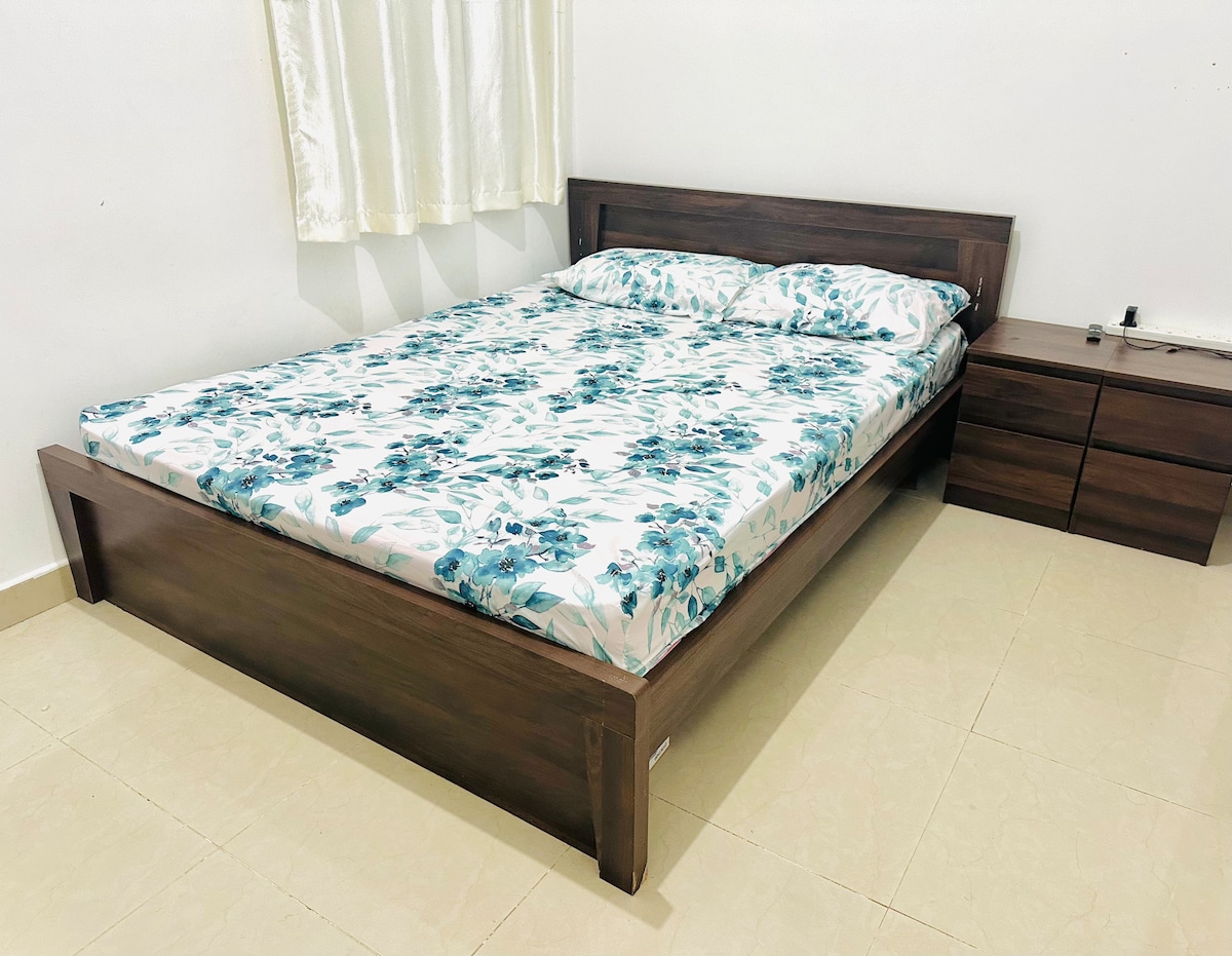 A spacious bed with a floral-patterned mattress is positioned against a simple, light-colored wall. A wooden nightstand with drawers accompanies the bed. Natural light filters through a nearby window, dressed with light curtains.