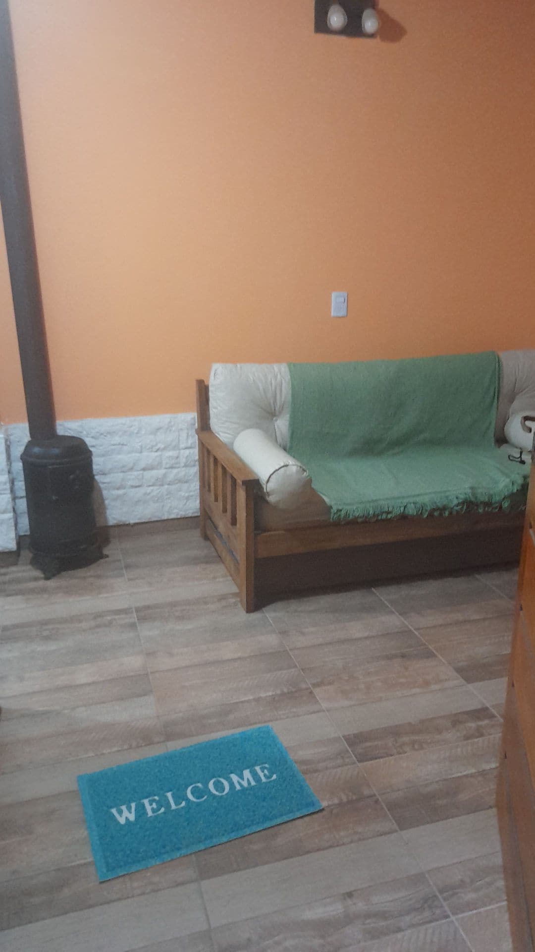 A comfortable seating area is highlighted by a wooden couch adorned with a green throw blanket. A small wood stove is positioned nearby, while a welcoming mat with the word 'WELCOME' can be seen on the tiled floor. The walls are painted a warm orange.