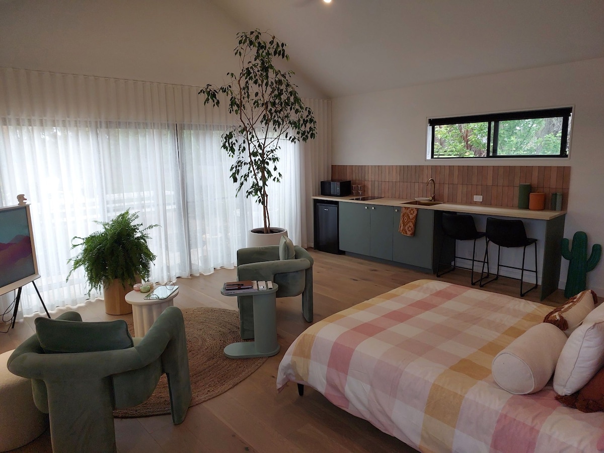 A modern open-plan space features a queen bed with a plaid blanket and ample natural light streaming through large windows with sheer curtains. A small kitchenette is visible, accompanied by a dining area with two bar stools. Relaxing seating is provided by two green armchairs.