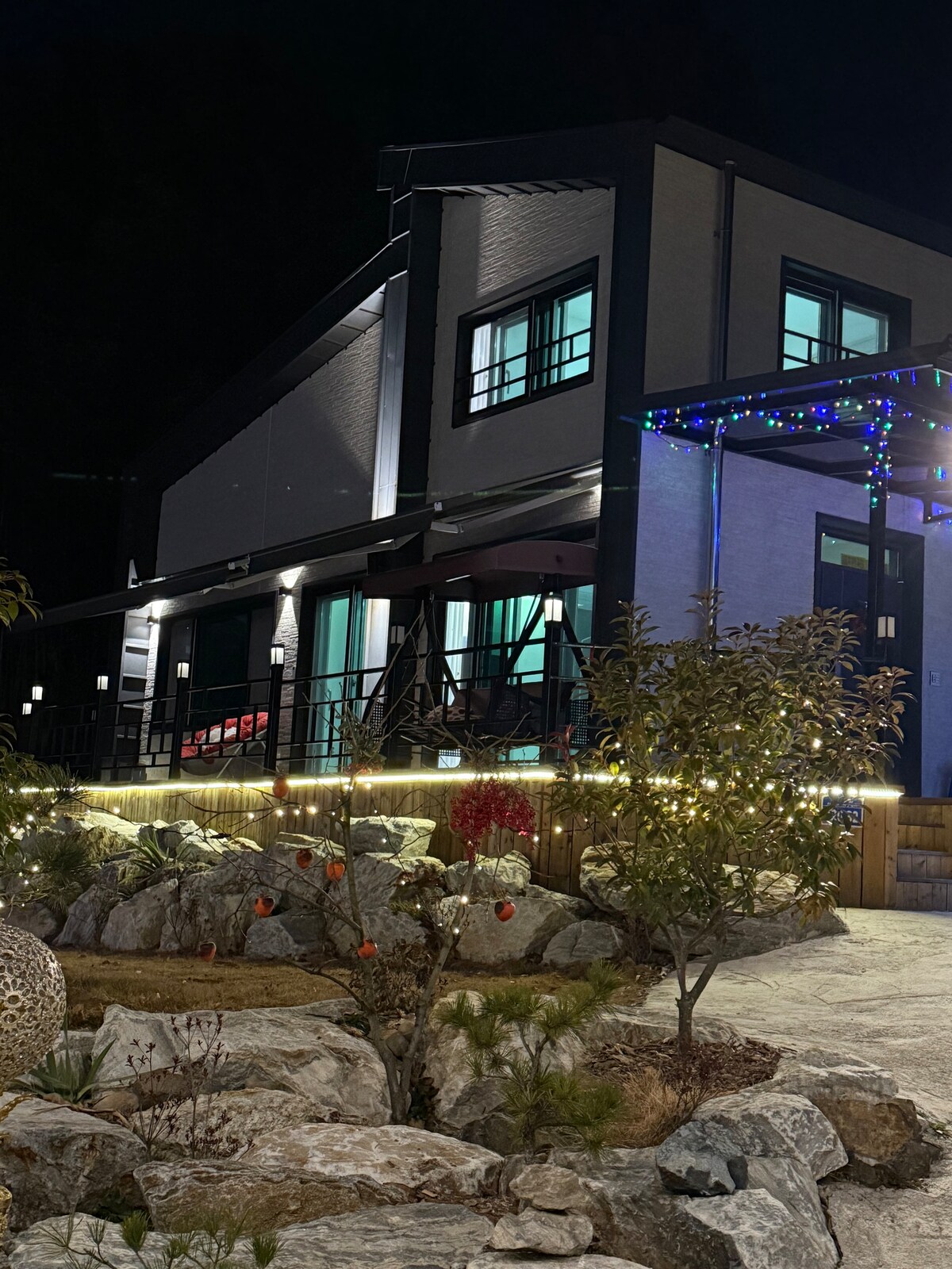 The two-story house is illuminated at night, showcasing its modern design. Soft lighting highlights the pathway and garden, with decorative lights adding a festive touch. The spacious balcony features seating, while large windows suggest a bright interior, contrasting against the dark sky.