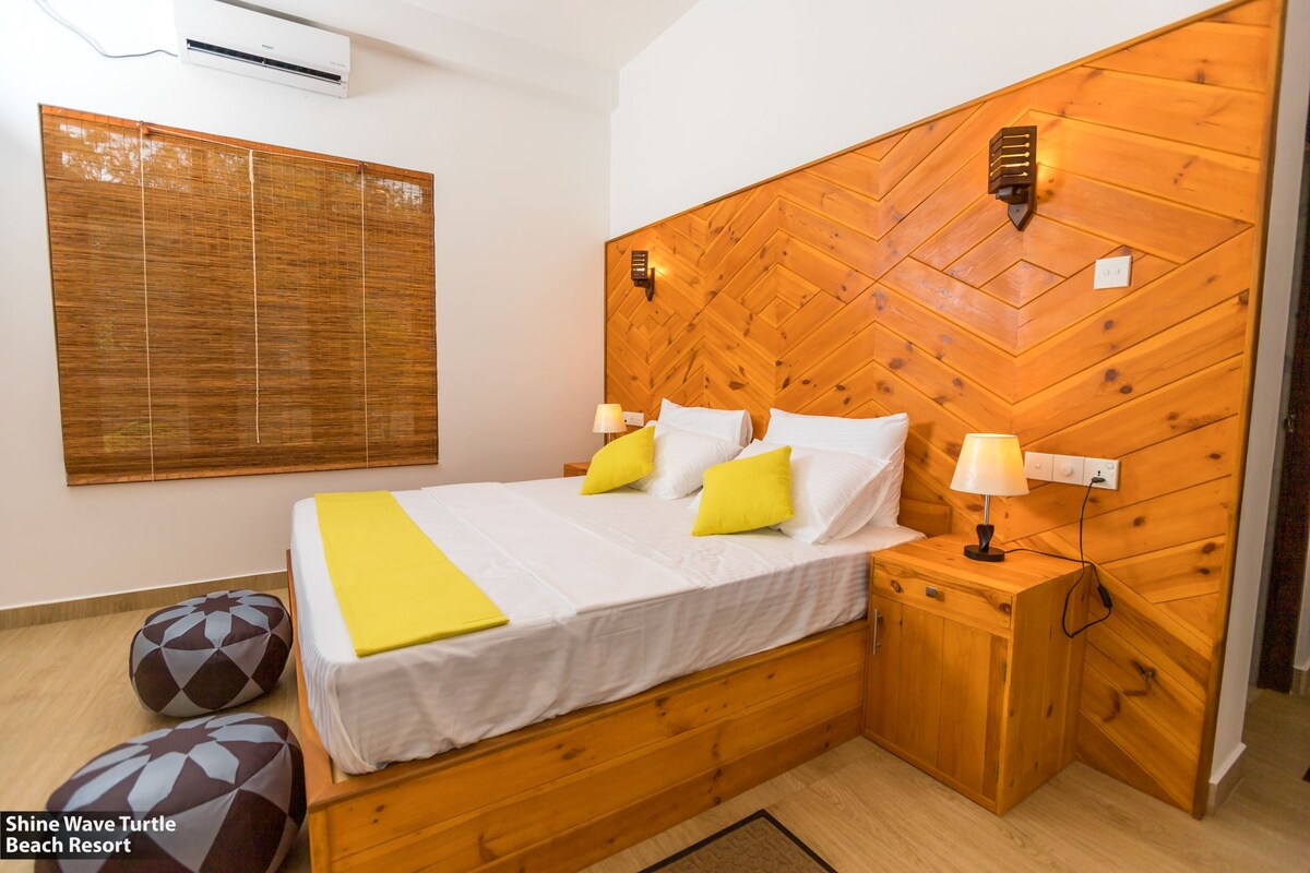 A cozy sleeping area features a wooden accent wall with a herringbone pattern. The spacious bed is dressed in crisp white linens with a yellow throw blanket. Two bedside lamps sit on matching wooden nightstands, complemented by round poufs positioned at the foot of the bed.