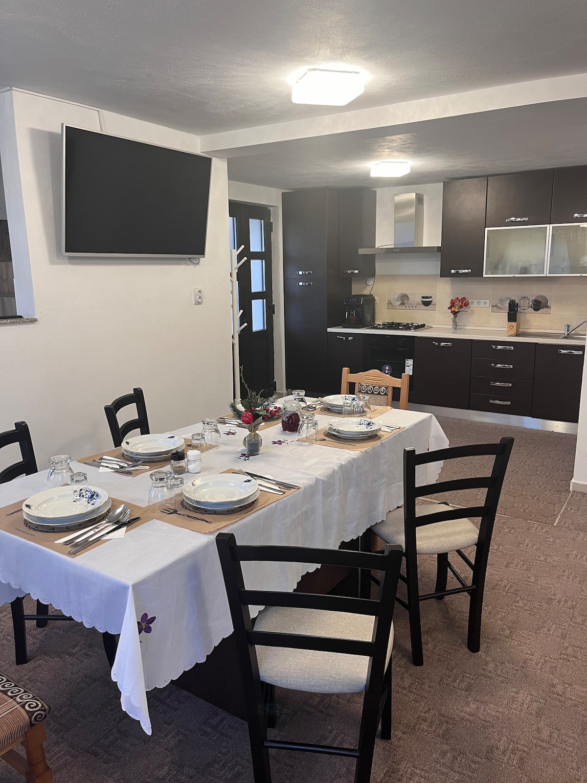 A dining area is presented with a table set for several guests, featuring elegant tableware and a simple floral centerpiece. The adjoining kitchen is equipped with modern appliances and cabinetry. A large wall-mounted TV is visible, enhancing the communal space.