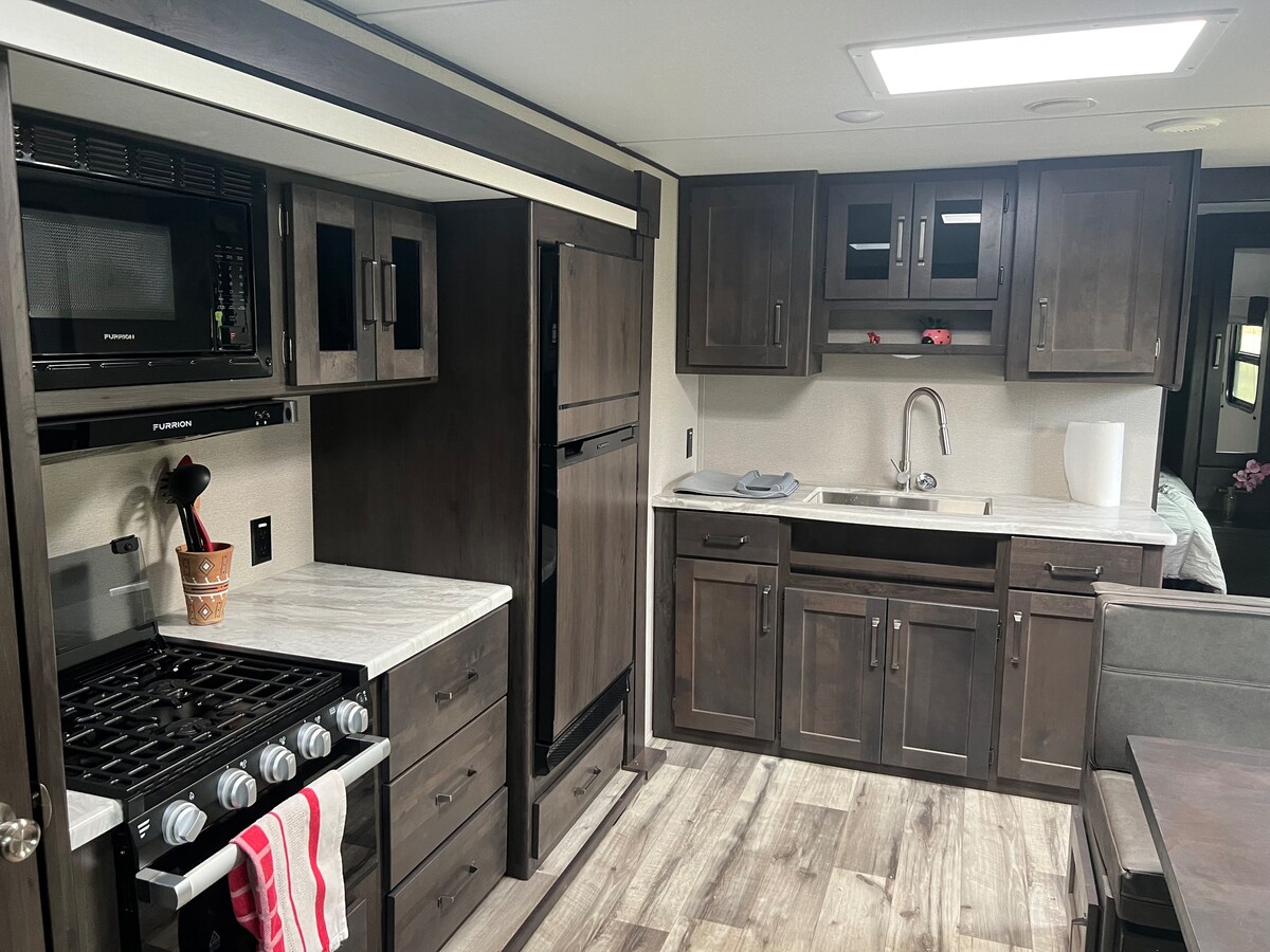 A modern kitchen is displayed, featuring dark cabinetry and stainless steel appliances. A gas stove with a visible red and white dish towel is positioned next to a sleek countertop. Ample storage is offered by cabinets and drawers, with a sink situated beneath a bright ceiling light.