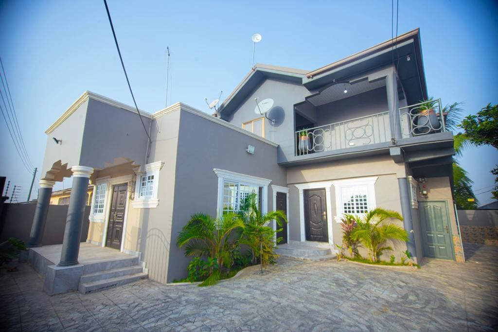 A modern two-story lodge exterior is highlighted, featuring a combination of gray walls and elegant architectural details. Lush greenery borders the entrance, and balconies are adorned with planters. The driveway leads to a welcoming atmosphere, showcasing a well-maintained facade with distinct doorways and windows.