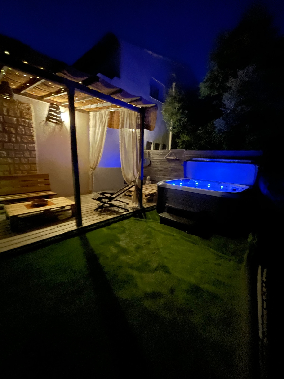 A serene outdoor space is illuminated by soft blue lights from a hot tub, with a wooden pergola draped in white fabric. Comfortable seating, including a bench and a reclining chair, is provided on the deck surrounded by well-maintained grass.
