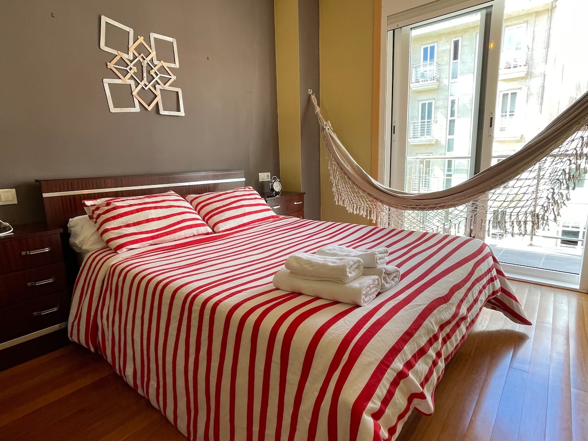 A spacious bedroom features a striped bedding set laid across the bed, with neatly folded white towels positioned at the foot. A hammock is attached to the walls, and large glass doors open to the outside, allowing natural light to fill the space.