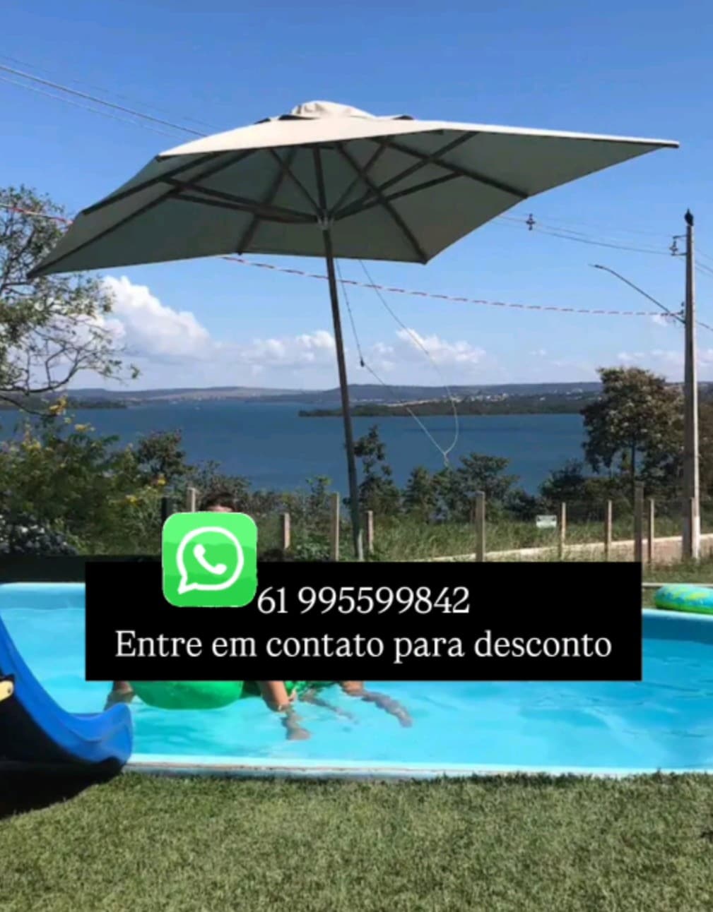 A serene pool area is visible, featuring a person relaxing beneath a large umbrella. The pool overlooks a calm lake, framed by green trees and grass. Clear blue water glistens under sunlight, inviting guests to enjoy the refreshing atmosphere.