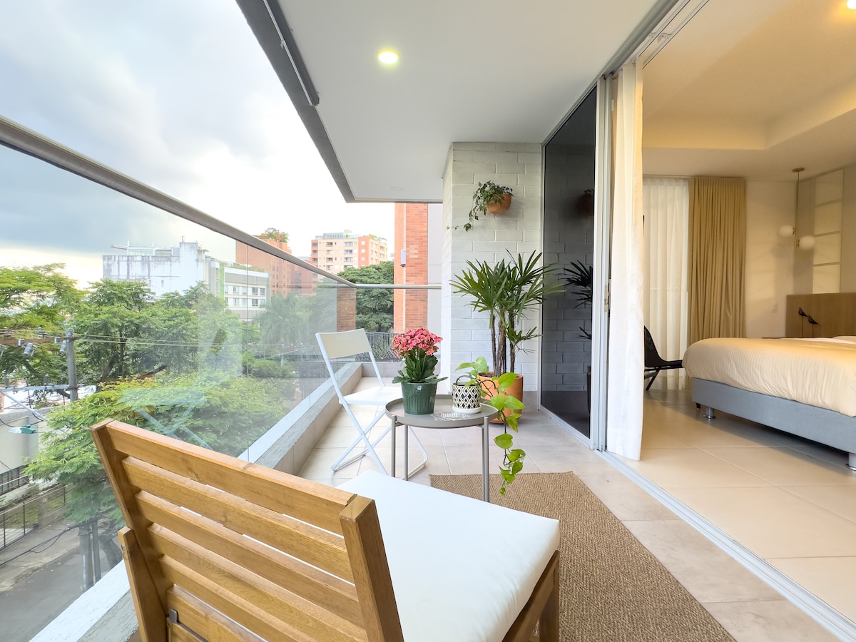 A private balcony is visible, featuring a wooden chair and small white table, adorned with potted plants and flowers. The view includes nearby buildings and greenery, while a spacious bed is seen through the glass doors, inviting natural light into the apartment.