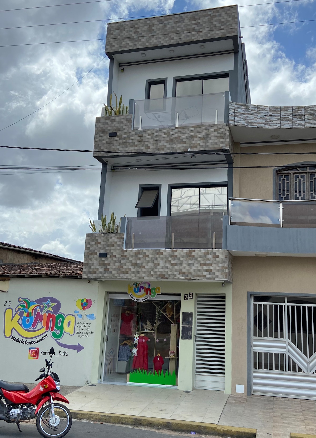 The building features a modern, three-story design with large windows and a balcony on the second floor. A colorful sign reading 'Koringa' is prominently displayed on the ground level, and the facade includes decorative elements that enhance its contemporary look.