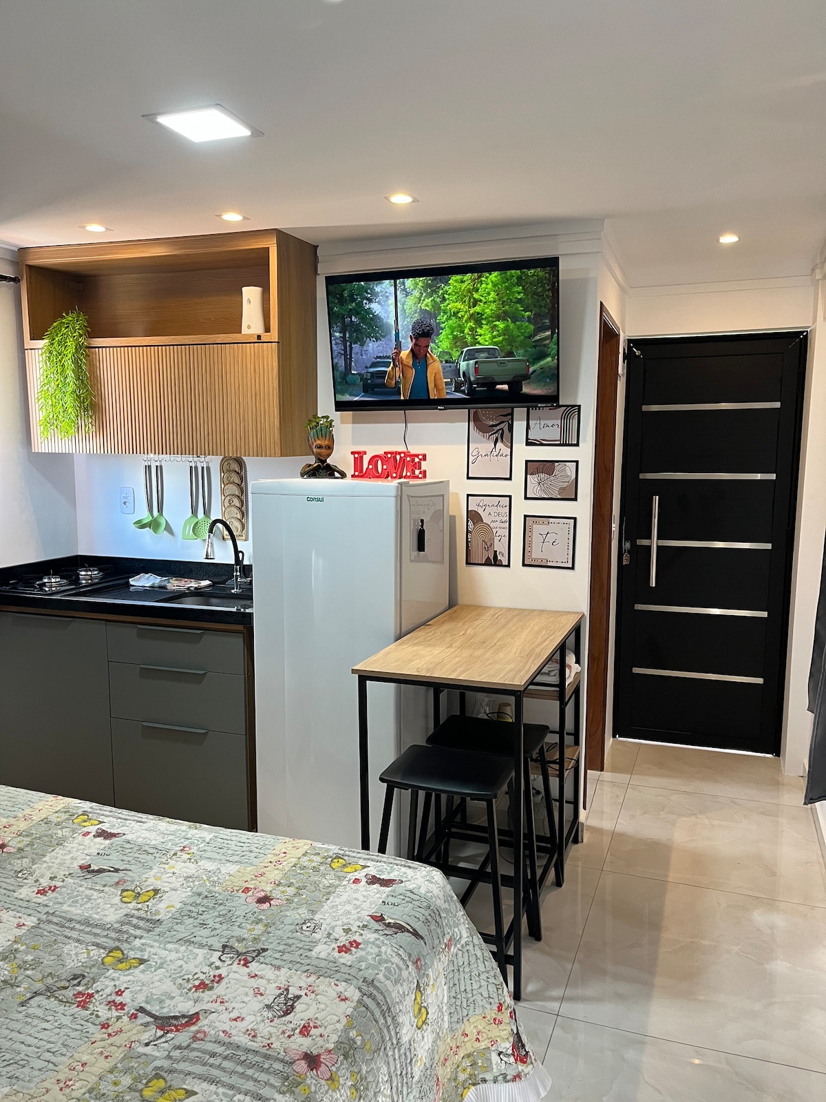 The compact living space features a kitchenette with modern appliances and a refrigerator. A small dining table with two chairs is positioned adjacent to artwork on the wall. A television is mounted above the kitchen area, providing entertainment options. Soft lighting enhances the cozy feel.