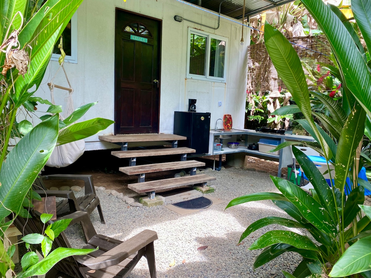 A cozy entrance is framed by lush greenery, with wooden steps leading up to a door. A small patio area features gravel flooring and is accompanied by simple wooden seating. Tropical plants and a hanging chair add to the serene outdoor environment.